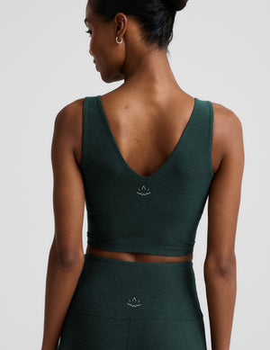 A woman modeling a dark forest green athletic set consisting of a V-neck crop tank and high-waisted pants, shot against a neutral light gray background.