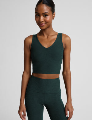 A woman modeling a dark forest green athletic set consisting of a V-neck crop tank and high-waisted pants, shot against a neutral light gray background.