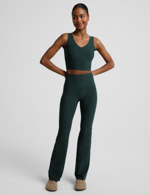 A woman modeling a dark forest green athletic set consisting of a V-neck crop tank and high-waisted pants, shot against a neutral light gray background.