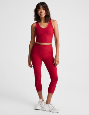 A woman modeling a matching red activewear set consisting of a sleeveless crop top and high-waisted capri leggings, paired with white sneakers, against a light gray studio background.