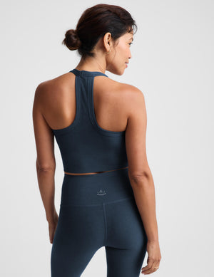 A woman modeling a matching slate blue activewear set, featuring a scoop-neck crop top and high-waisted leggings, against a light gray studio background.