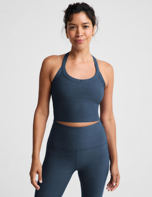 A woman modeling a matching slate blue activewear set, featuring a scoop-neck crop top and high-waisted leggings, against a light gray studio background.