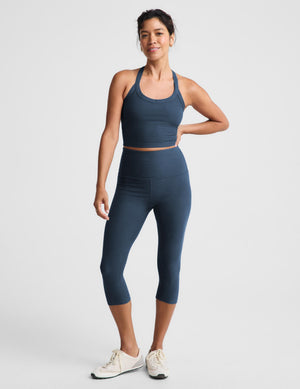 A woman modeling a matching slate blue activewear set, featuring a scoop-neck crop top and high-waisted leggings, against a light gray studio background.