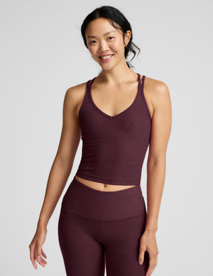 A woman modeling a matching athletic set consisting of a dark plum-burgundy V-neck crop tank top and high-waisted leggings, photographed against a solid light gray studio background.