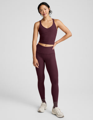 A woman modeling a matching athletic set consisting of a dark plum-burgundy crop top and high-waisted leggings, paired with light gray sneakers, against a light gray studio background.