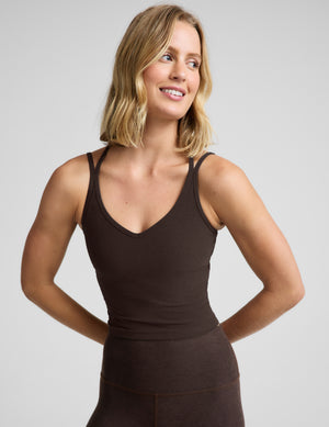 Woman wearing a brown midway length tank with crossover back straps and brown leggings against a plain studio background. 