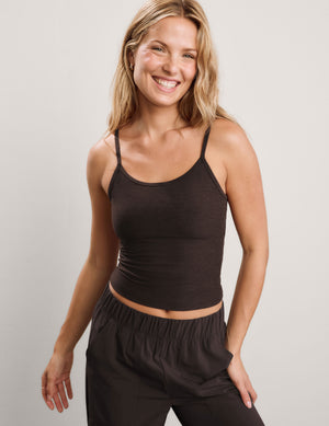 A smiling blonde woman wearing a dark brown spaghetti-strap tank top and matching dark brown pants, photographed against a plain light gray studio background.