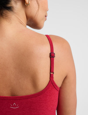 Close-up back view of a woman wearing a heathered red tank top with adjustable  straps and a small white logo near the lower left shoulder on a light gray background.
