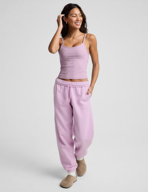 A woman modeling a matching light purple/lavender lounge set consisting of a spaghetti-strap camisole top and relaxed sweatpants, paired with beige slip-on shoes, against a solid light gray background.