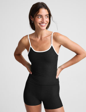 A woman wearing a black camisole tank top with white piping and black biker shorts against a plain studio background. 