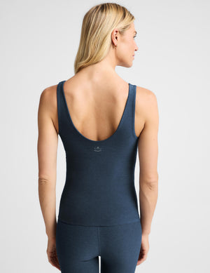 A woman shown from the back wearing a dark slate blue athletic tank top and matching leggings against a light gray studio background.