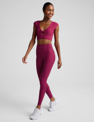 A woman modeling a matching athletic set featuring a short-sleeve crop top with a twisted front detail and high-waisted leggings, in a heathered magenta-pink color, against a solid light gray background.