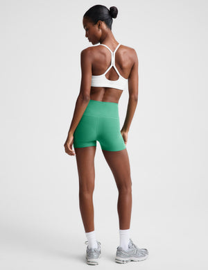 A woman modeling high-waisted green athletic shorts, paired with a white sports bra, white socks, and gray sneakers, against a light gray studio background. 