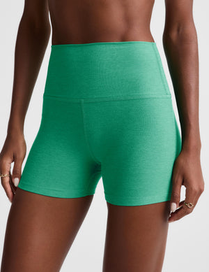 A close-up of a model wearing high-waisted athletic shorts in a heathered green color, showing the front and side fit against a light gray studio background.