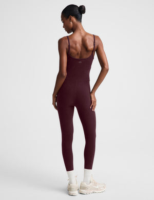 A model wearing a burgundy midi length tank jumpsuit, styled with white socks and beige sneakers, against a light gray studio background.