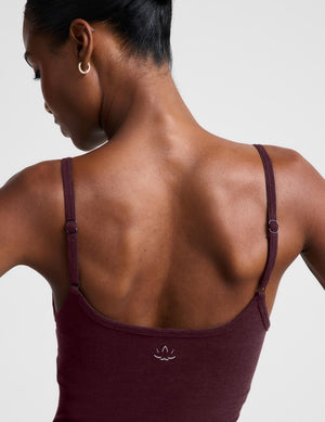 A model wearing a burgundy midi length tank jumpsuit, styled with white socks and beige sneakers, against a light gray studio background.