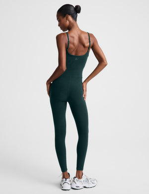 A woman modeling a dark forest green sleeveless bodysuit, paired with white sneakers, against a light gray studio background.