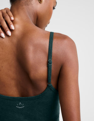 Close-up back view of a woman wearing a dark green midi jumpsuit with thin adjustable shoulder straps and a small white lotus logo near the lower left back, against a light gray studio background.
