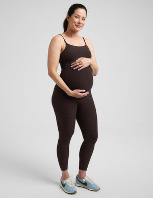 Pregnant woman wearing a brown midi length jumpsuit against a plain background. 