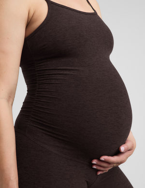 Pregnant woman wearing a brown midi length jumpsuit against a plain background. 