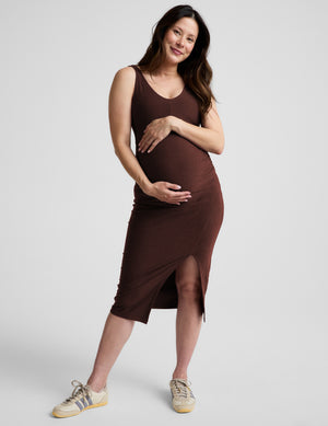 Pregnant woman wearing a brown maternity midi length dress with a front slit against a plain background. 