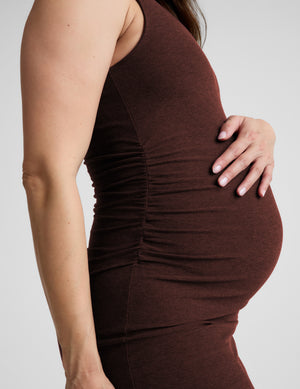 Pregnant woman wearing a brown maternity midi length dress with a front slit against a plain background. 