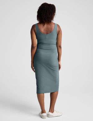 Pregnant woman wearing a green maternity midi length tank dress with a front side slit against a plain background. 
