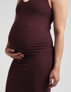 Pregnant woman wearing a red maternity dress and brown loafers against a plain studio background. 
