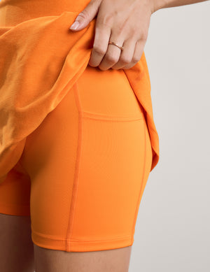 A woman modeling a bright orange sleeveless mini dress with white sneakers against a neutral studio backdrop.