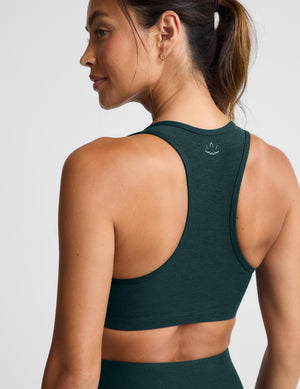 A woman modeling a dark forest green athletic sports bra and matching high-waisted leggings against a light gray studio background.