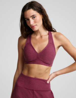A woman modeling a heathered burgundy sports bra and matching high-waisted leggings against a light gray studio background.