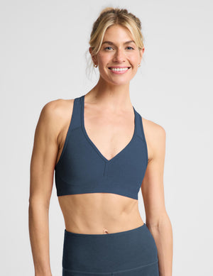 A smiling blonde woman wearing a dark slate blue sports bra and matching high-waisted leggings, photographed against a solid light gray studio background.
