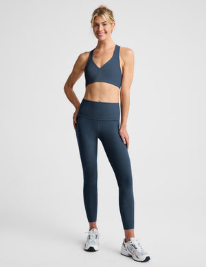 A woman modeling a matching athletic set consisting of a navy blue sports bra and high-waisted leggings, paired with white and gray sneakers, against a light gray studio background.
