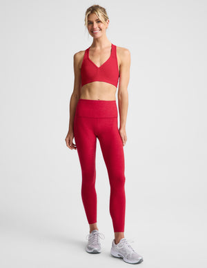 A woman modeling a bright red sports bra and matching red leggings against a light gray studio background.