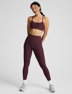 A woman modeling a matching athletic set consisting of a scoop-neck sports bra and high-waisted leggings in a deep red color, shot against a solid light gray background.