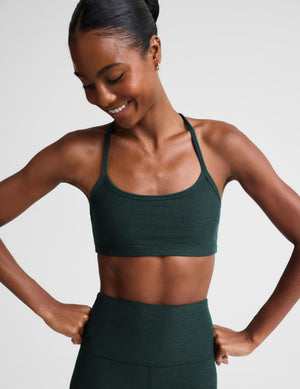A woman wearing a dark forest green athletic sports bra and matching high-waisted pants, posing with hands on hips against a light gray studio background.