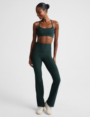 A woman modeling a matching dark green activewear set consisting of a sports bra and high-waisted bootcut leggings, paired with white sneakers, against a light gray studio background.