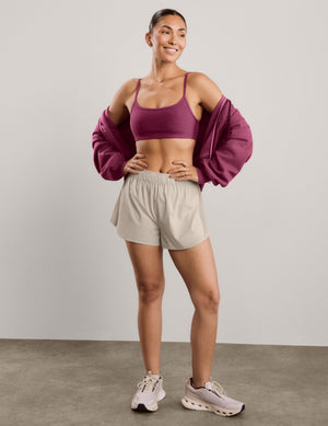 A woman modeling a heathered red purple sports bra and light beige athletic shorts against a solid light gray studio background.