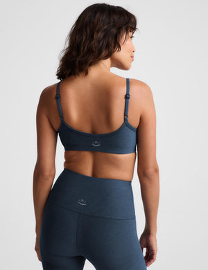 A woman modeling a dark slate blue sports bra and matching high-waisted leggings against a light gray studio background.