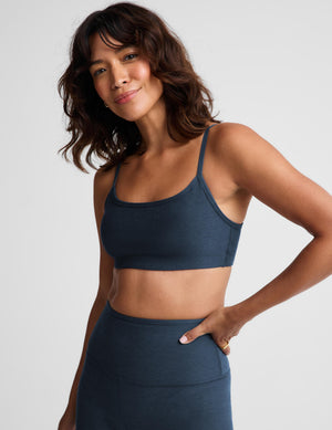 A woman modeling a dark slate blue sports bra and matching high-waisted leggings against a light gray studio background.