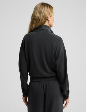 A woman shown from the back wearing a black long-sleeve mock neck top and matching black pants against a light gray studio background.