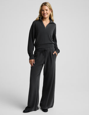 A woman modeling a black long-sleeve quarter-zip pullover and matching black wide-leg drawstring pants against a light gray studio background.