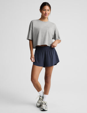 A woman modeling a light gray cropped short-sleeve t-shirt and navy blue athletic shorts against a light gray studio background.