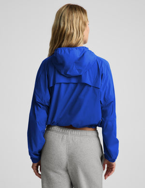 A woman shown from the back wearing a bright royal blue cropped hooded windbreaker jacket and light gray sweatpants against a light gray studio background.
