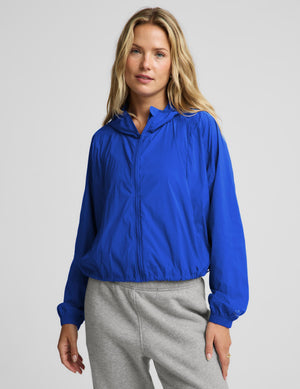 A woman wearing a bright royal blue lightweight zip-up hooded jacket and light gray sweatpants, standing against a plain light gray studio background.