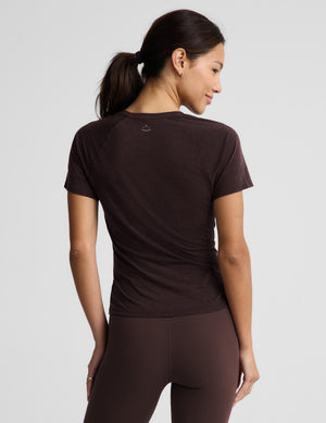 A woman shown from the back wearing a fitted short-sleeve athletic top and matching leggings in a dark brown color, against a light gray studio background.