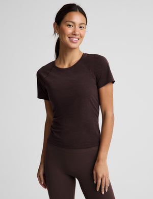A woman wearing a brown active short sleeve t-shirt and brown leggings against a light gray studio background. 