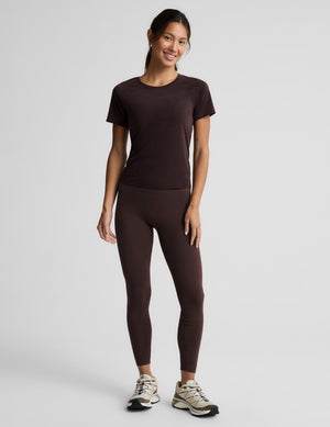 A woman modeling a dark brown short-sleeve athletic t-shirt and matching dark brown high-waisted leggings, paired with beige and black running shoes, against a light gray studio background.
