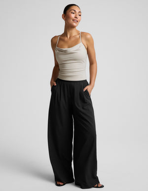 Woman wearing a beige cowl neck tank top and black trousers against a plain background. 