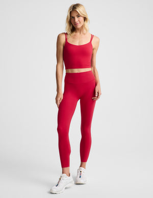 A woman modeling a matching red activewear set consisting of a spaghetti-strap crop top and high-waisted leggings, paired with white sneakers, against a light gray studio background.